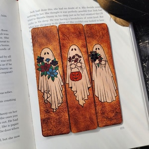 May include: Three wooden bookmarks with ghost illustrations. Each ghost is draped in a white sheet and holding flowers or a pumpkin. The bookmarks are on a book page with text.