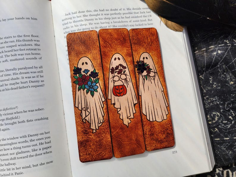 May include: Three wooden bookmarks with ghost illustrations. Each ghost is draped in a white sheet and holding flowers or a pumpkin. The bookmarks are on a book page with text.