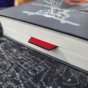 May include: Close-up of a black hardback book with a red ribbon bookmark. The book cover features a detailed illustration of a dragonfly.