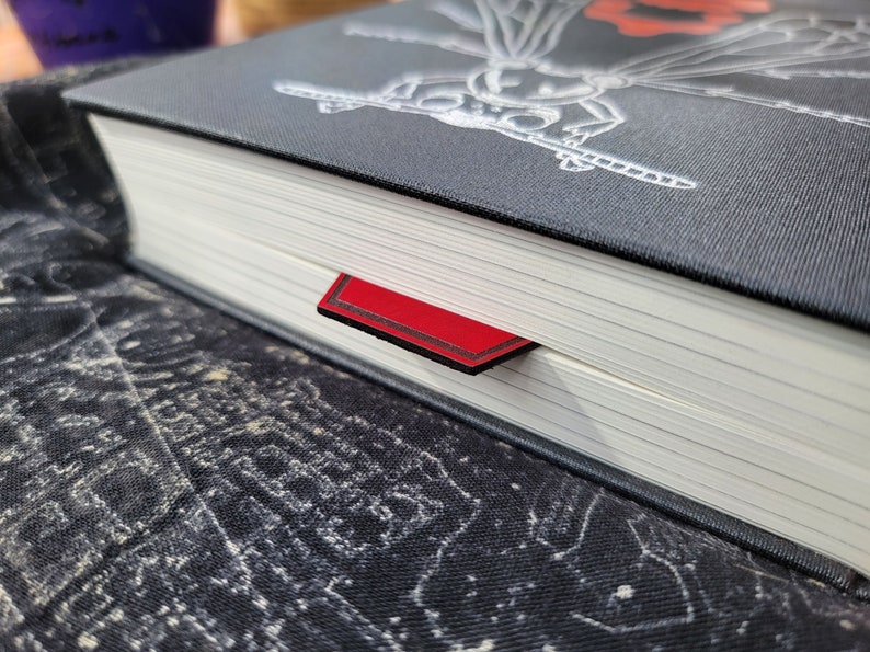May include: Close-up of a black hardback book with a red ribbon bookmark. The book cover features a detailed illustration of a dragonfly.