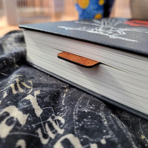 May include: A hardcover book with a black cover and a brown leather bookmark. The book's pages are white, and the bookmark is inserted between them. The book is resting on a dark patterned fabric.