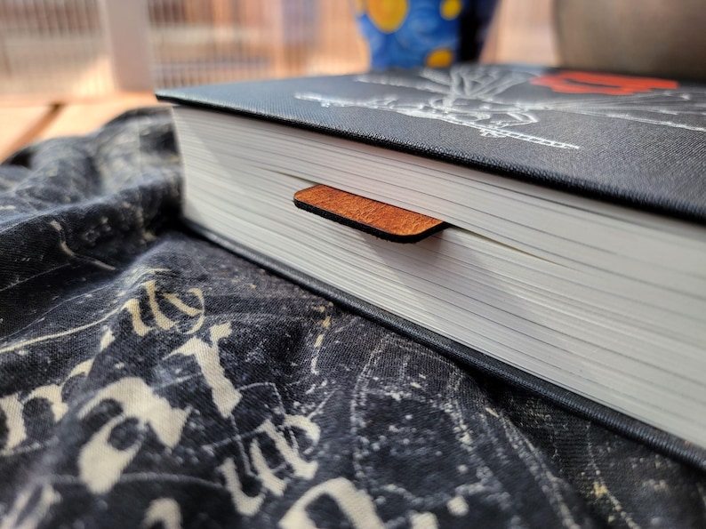 May include: A hardcover book with a black cover and a brown leather bookmark. The book's pages are white, and the bookmark is inserted between them. The book is resting on a dark patterned fabric.
