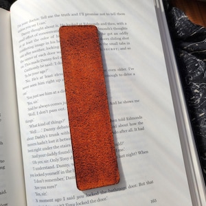 May include: A rectangular, textured, orange leather bookmark is placed on an open book. The bookmark is a solid color with a slightly rough surface. The book pages are white with black text.