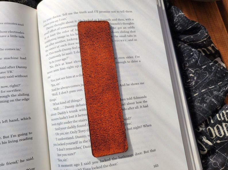 May include: A rectangular, textured, orange leather bookmark is placed on an open book. The bookmark is a solid color with a slightly rough surface. The book pages are white with black text.