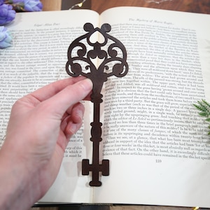 May include: A dark brown wooden key-shaped bookmark with intricate details and a heart-shaped cutout. The bookmark is being held in a hand against a page of a book.