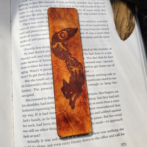 May include: A brown leather bookmark with a black silhouette of a fox running through a forest scene. The bookmark has a moon and stars design at the top.