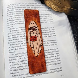 May include: A Halloween-themed bookmark with a ghost illustration holding flowers and a jack-o'-lantern. The bookmark is rectangular with a textured, brown leather-like appearance, and is placed on an open book.