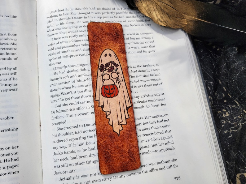 May include: A Halloween-themed bookmark with a ghost illustration holding flowers and a jack-o'-lantern. The bookmark is rectangular with a textured, brown leather-like appearance, and is placed on an open book.