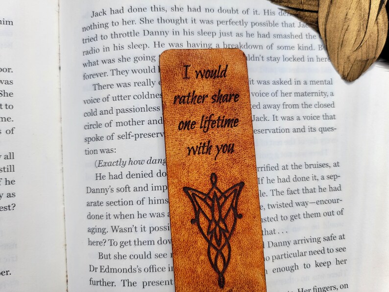 May include: A brown leather bookmark with a wood-burned design of the One Ring from The Lord of the Rings. The bookmark has the text "I would rather share one lifetime with you" inscribed on it.