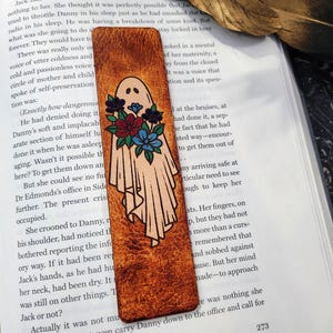 May include: Leather bookmark featuring a ghost design with a floral bouquet. The ghost is in a flowing sheet with a simple face. The flowers are red, blue, and teal. The bookmark is rectangular and has a textured, brown surface.