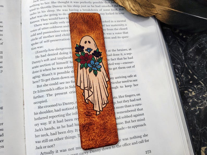 May include: Leather bookmark featuring a ghost design with a floral bouquet. The ghost is in a flowing sheet with a simple face. The flowers are red, blue, and teal. The bookmark is rectangular and has a textured, brown surface.