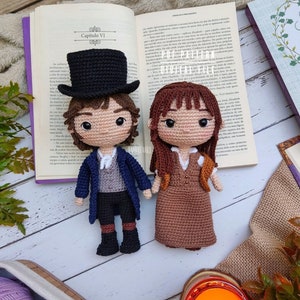 Op de afbeelding: Twee gehaakte amigurumi poppen, een met een zwarte hoge hoed en een blauw jasje, de andere met bruin haar en een bruine jurk. De poppen zitten op een boek met de tekst "Capítulo VI" zichtbaar.