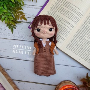 Może przedstawiać: Lalka szydełkowa z brązowymi włosami, brązową sukienką i brązową kurtką. Lalka siedzi na książce z portugalskim tekstem. Książka jest otwarta na stronie z tytułem "Capítulo IV".