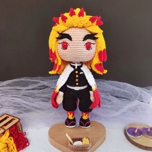 Puede incluir: Muñeca de crochet de un personaje de anime con cabello rojo y amarillo, ropa negra y blanca y ojos rojos. La muñeca está de pie sobre una base de madera con un pequeño tazón de comida.