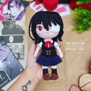 May include: Crochet amigurumi doll of a girl with black hair, a white eye patch, and a red eye. She is wearing a black and blue school uniform with a red bow. The doll is holding a PDF pattern for a crochet doll.