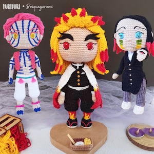 Puede incluir: Tres personajes de anime de crochet, uno con cabello rojo y amarillo, otro con cabello rosado y otro con cabello negro. Los personajes están de pie sobre una superficie blanca con una base de madera en forma de corazón y una pequeña caja de madera con palillos y comida.