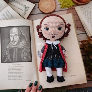 Puede incluir: Una muñeca de crochet de William Shakespeare, vestida con un atuendo rojo y azul, sosteniendo una pluma. La muñeca está sentada en un libro abierto con un retrato de Shakespeare en la página.