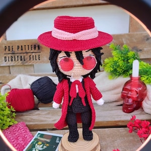 Puede incluir: Muñeca amigurumi de crochet de un personaje que lleva un sombrero rojo, una chaqueta roja y pantalones negros. La muñeca tiene el pelo negro, gafas rojas y una corbata de lazo roja. La muñeca está de pie sobre una base de madera. La imagen incluye el texto "PDF PATTERN DIGITAL FILE".