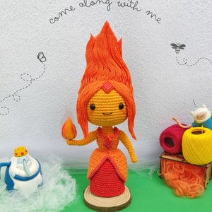 Peut inclure: Poupée en crochet de la Princesse de Feu du dessin animé Adventure Time. La poupée porte une robe rouge et a les cheveux orange. La poupée tient une petite flamme orange dans sa main droite. La poupée est debout sur une base en bois. L'arrière-plan est une surface verte avec un mur blanc et de la laine.
