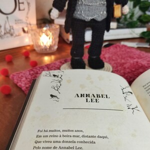 May include: A crocheted doll wearing a black suit with a gray vest and a red tie. The doll is holding a book with the title "Annabel Lee" written on the cover. The book is open to a page with a poem about a woman named Annabel Lee.