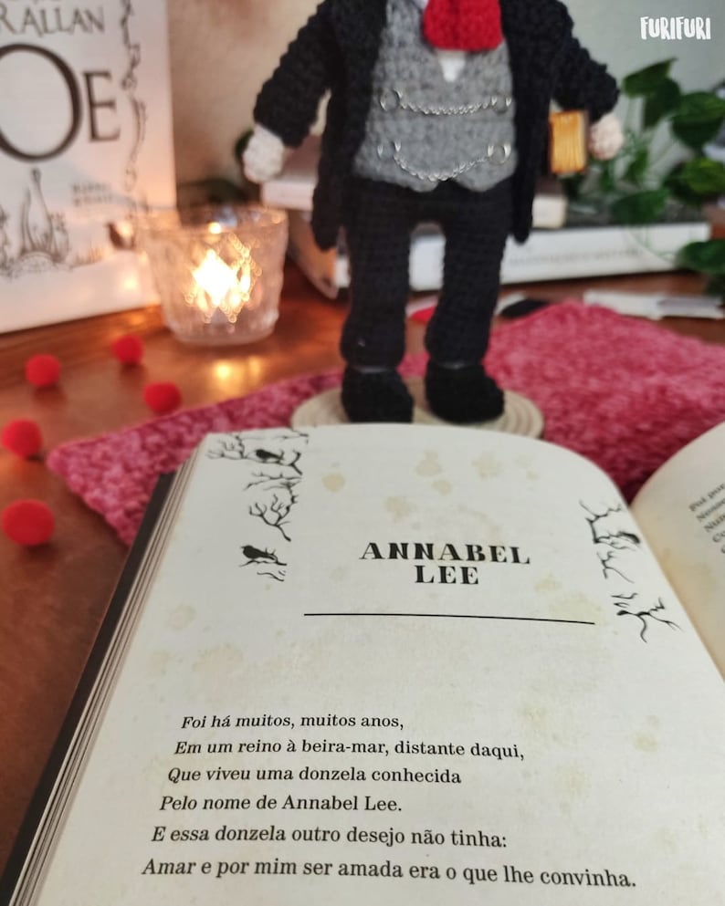 May include: A crocheted doll wearing a black suit with a gray vest and a red tie. The doll is holding a book with the title "Annabel Lee" written on the cover. The book is open to a page with a poem about a woman named Annabel Lee.