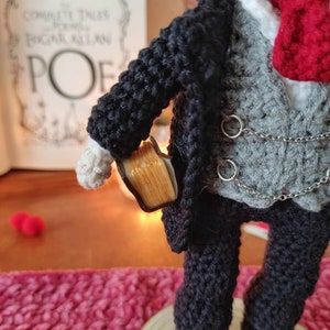 May include: Crocheted doll wearing a black suit with a gray vest, a white shirt, and a red bow tie. The doll is holding a small book in its right hand.