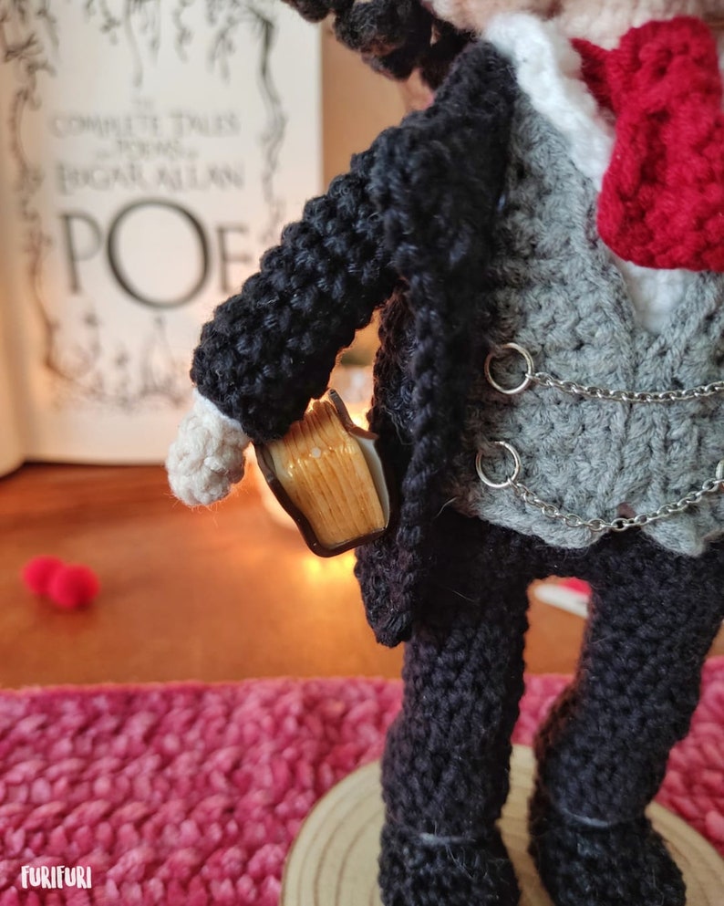 May include: Crocheted doll wearing a black suit with a gray vest, a white shirt, and a red bow tie. The doll is holding a small book in its right hand.