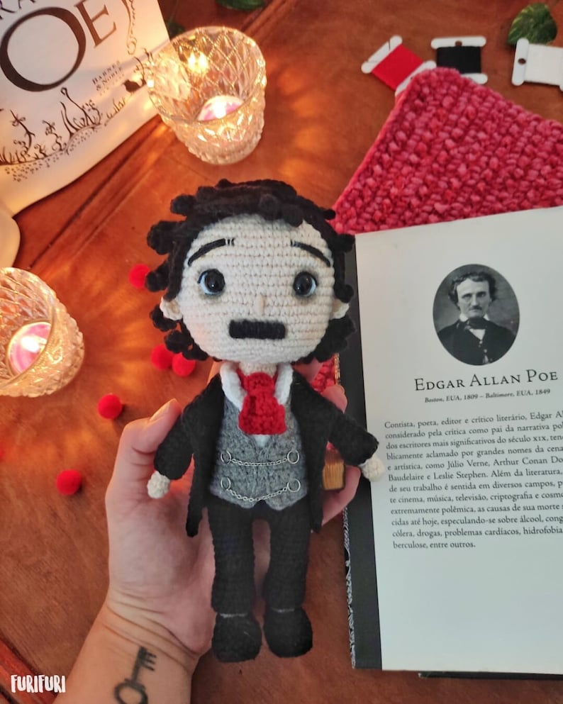 May include: Crocheted amigurumi doll of Edgar Allan Poe, wearing a black suit, gray vest, and red tie. The doll is holding a book with the text "Edgar Allan Poe" on the cover. The book is open to a page with a biography of Poe.