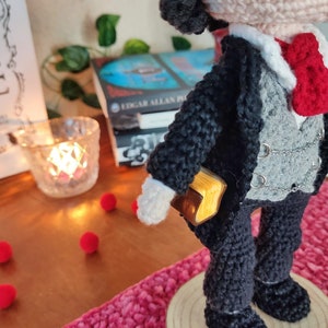 May include: Crocheted amigurumi doll with black hair, wearing a black suit with a gray vest, a white shirt, a red bow tie, and a gold book tucked into the pocket. The doll is standing on a wooden base.