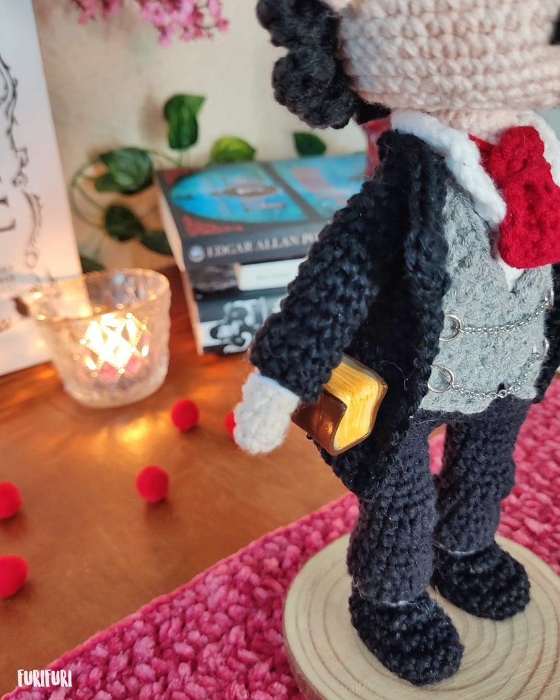 May include: Crocheted amigurumi doll with black hair, wearing a black suit with a gray vest, a white shirt, a red bow tie, and a gold book tucked into the pocket. The doll is standing on a wooden base.
