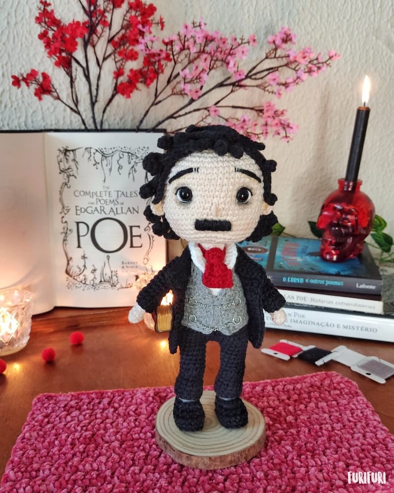 May include: Crocheted amigurumi doll of Edgar Allan Poe holding a book. The doll is wearing a black suit, a white shirt, a red tie, and a gray vest. The doll is standing on a wooden base. The doll is in front of a book titled "The Complete Tales and Poems of Edgar Allan Poe".