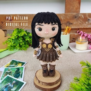 Może przedstawiać: Wzór na amigurumową lalkę wojowniczki z czarnymi włosami, brązową skórzaną zbroją i złotymi akcentami. Wzór jest dostępny jako cyfrowy plik PDF.