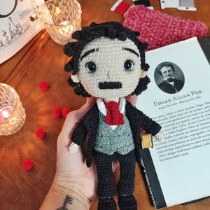 May include: Crocheted amigurumi doll of Edgar Allan Poe, wearing a black suit, gray vest, and red tie. The doll is holding a small book in its right hand. The doll is sitting on a book with the title "Edgar Allan Poe" on the cover.