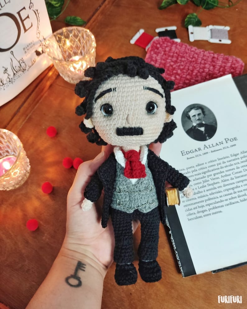 May include: Crocheted amigurumi doll of Edgar Allan Poe, wearing a black suit, gray vest, and red tie. The doll is holding a small book in its right hand. The doll is sitting on a book with the title "Edgar Allan Poe" on the cover.