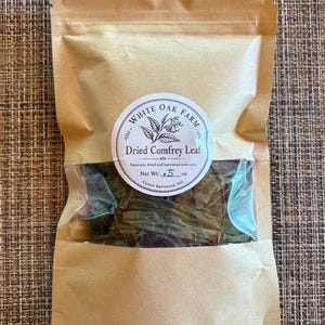 May include: A brown paper pouch with a clear window containing dark green dried comfrey leaves. A circular white label features the text White Oak Farm, Dried Comfrey Leaf, and Net Wt: .5 oz, with a botanical illustration of leaves.