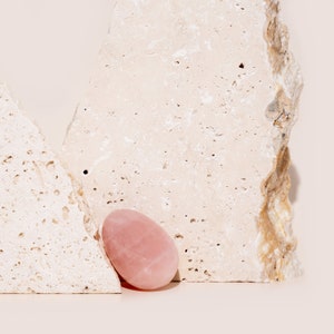 May include: A light pink rose quartz egg-shaped crystal resting on a light beige textured surface.