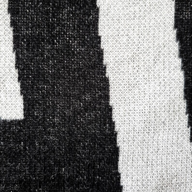 K&ouml;nnte beinhalten: Nahaufnahme eines Strickstoffs mit einem auff&auml;lligen schwarz-wei&szlig; gestreiften Muster. Die Textur des Stricks ist sichtbar, mit einer Mischung aus schwarzen und wei&szlig;en F&auml;den, die das Design bilden. Die Streifen sind vertikal und gleichm&auml;&szlig;ig angeordnet.