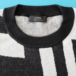 K&ouml;nnte beinhalten: Nahaufnahme eines X-Large-Sweaters mit einem schwarz-wei&szlig;en geometrischen Muster. Der Pullover hat einen schwarzen gerippten Rundhalsausschnitt und das Etikett lautet "X-LARGE MADE IN CHINA". Der Pullover ist aus einem weichen, strukturierten Strickstoff gefertigt.
