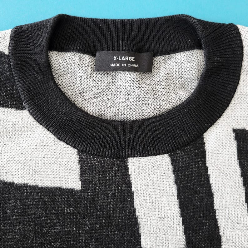 K&ouml;nnte beinhalten: Nahaufnahme eines X-Large-Sweaters mit einem schwarz-wei&szlig;en geometrischen Muster. Der Pullover hat einen schwarzen gerippten Rundhalsausschnitt und das Etikett lautet "X-LARGE MADE IN CHINA". Der Pullover ist aus einem weichen, strukturierten Strickstoff gefertigt.