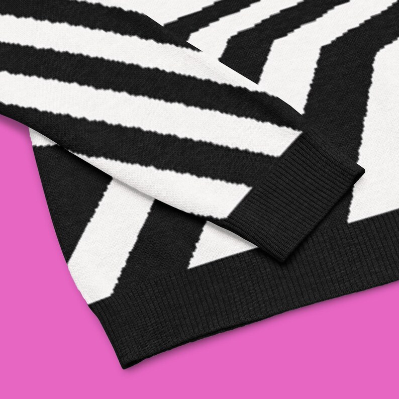 K&ouml;nnte beinhalten: Nahaufnahme eines schwarz-wei&szlig; gestreiften Pullovers mit einem auff&auml;lligen geometrischen Muster. Der Pullover hat dicke, diagonale Streifen und eine gerippte schwarze Manschette. Der Saum ist ebenfalls schwarz. Das Kleidungsst&uuml;ck ist aus weichem Material.