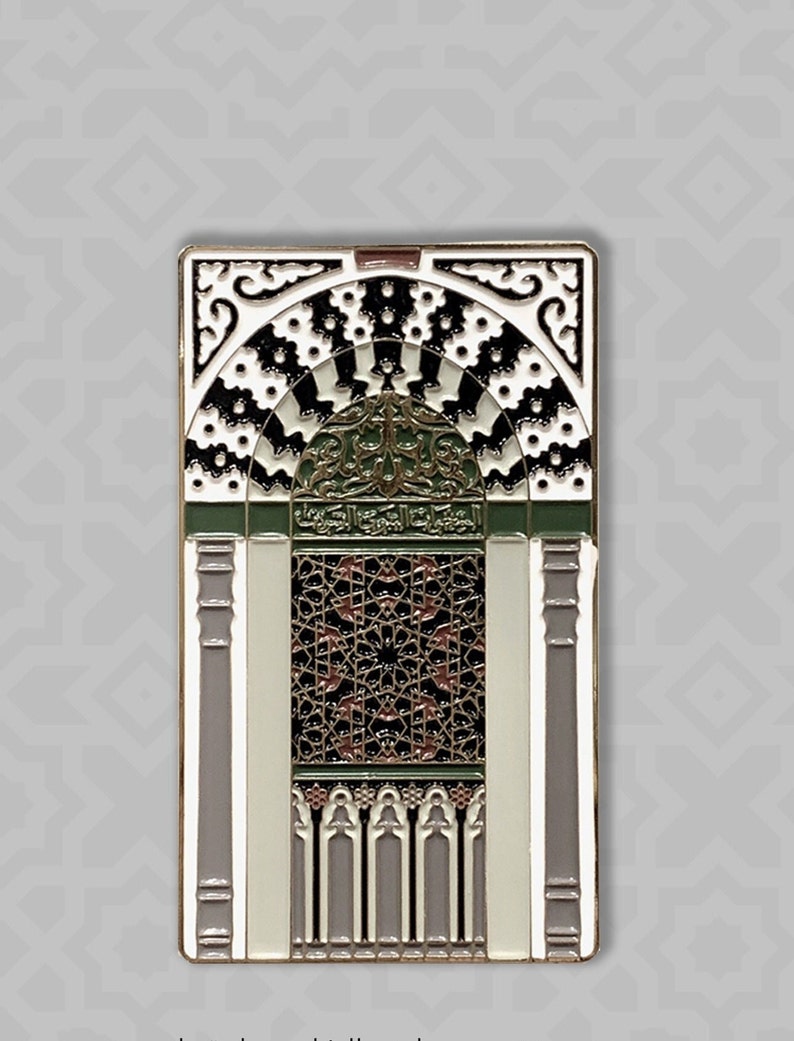 May include: A gold-toned enamel pin depicting an intricate archway with black and white geometric patterns, green and brown accents, and Arabic calligraphy.