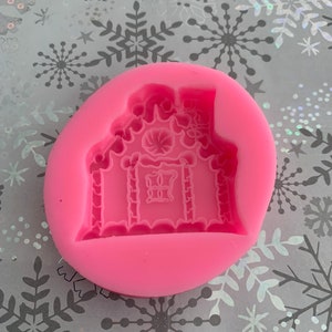 May include: Pink silicone mold for making gingerbread house shaped cookies. The mold has a detailed design with a candy cane roof and a window.