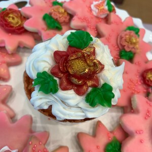 May include: A white cupcake with red and green frosting and a gold accent. The cupcake is surrounded by pink star-shaped cookies with gold accents.