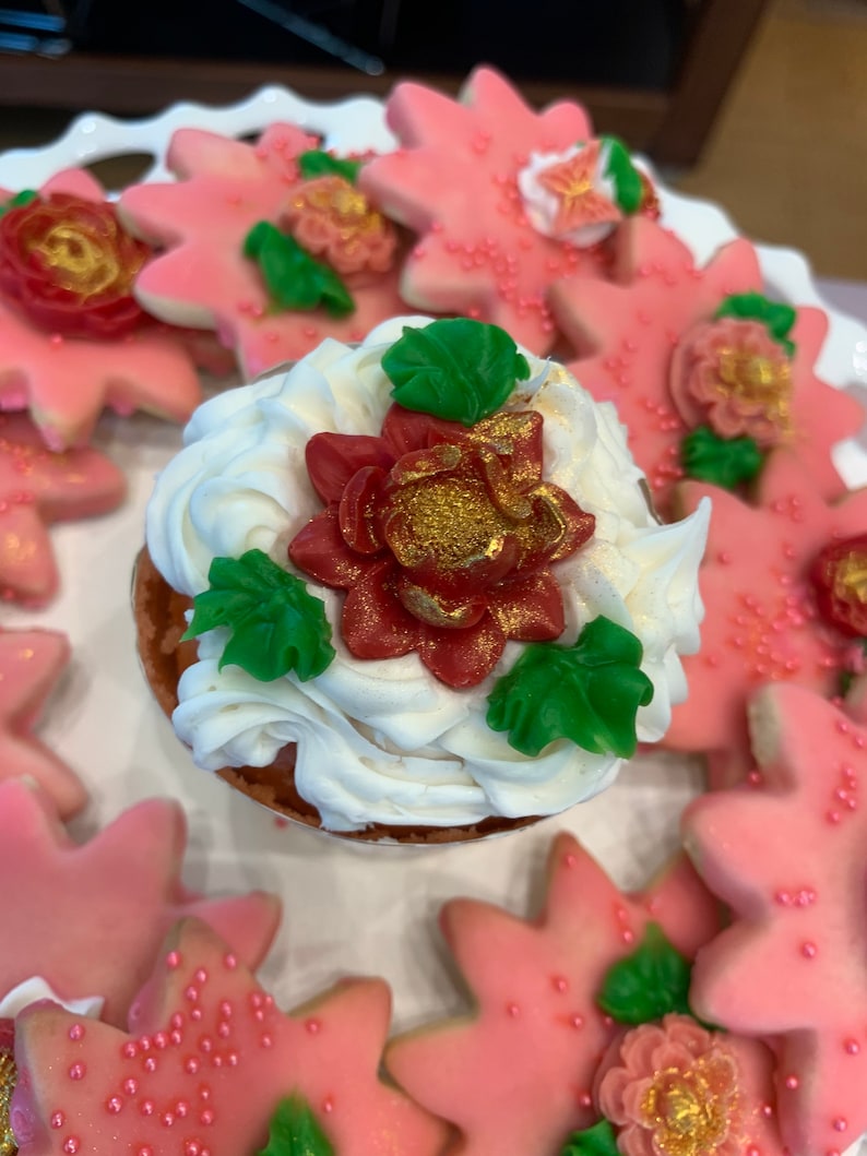 May include: A white cupcake with red and green frosting and a gold accent. The cupcake is surrounded by pink star-shaped cookies with gold accents.