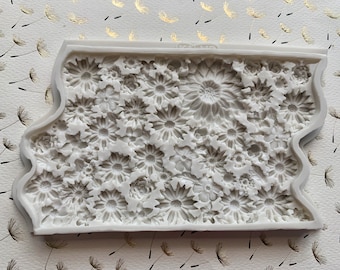 May include: A white silicone mold featuring a detailed floral design. The mold is rectangular with a decorative edge and filled with various sizes of daisy-like flowers. The background is a white paper with gold dandelion accents.