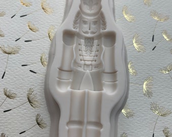 May include: A white silicone mold for making a nutcracker figurine. The mold is shaped like a standing nutcracker with a hat, mustache, and a detailed uniform.
