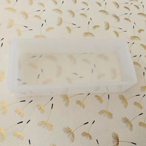 May include: A rectangular, translucent silicone mold. The mold is empty and sits on a cream-colored surface decorated with a repeating pattern of gold dandelion seed heads and black stems. The mold is likely for crafting.