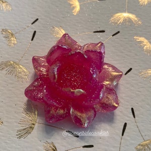 May include: A pink sugar flower with gold glitter accents. The flower is on a white background with gold dandelion designs.