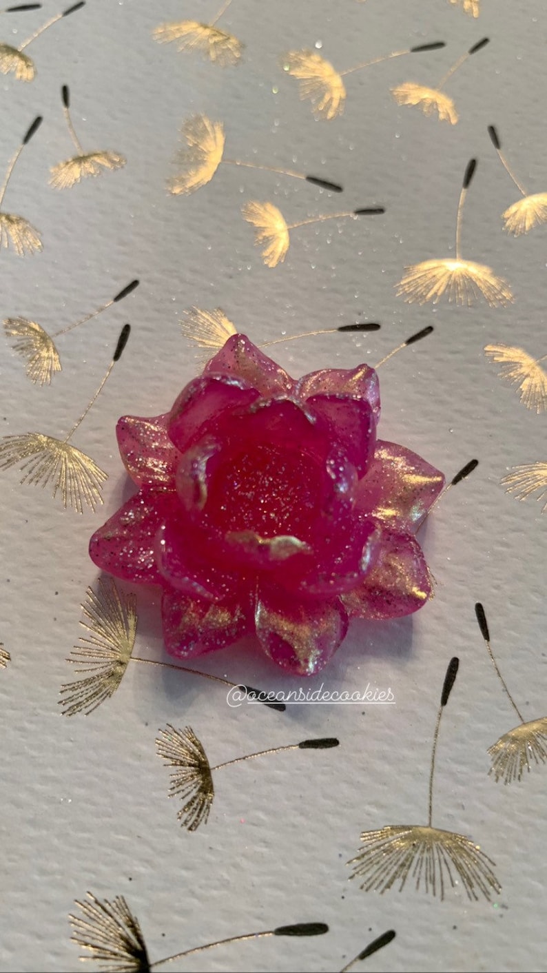 May include: A pink sugar flower with gold glitter accents. The flower is on a white background with gold dandelion designs.