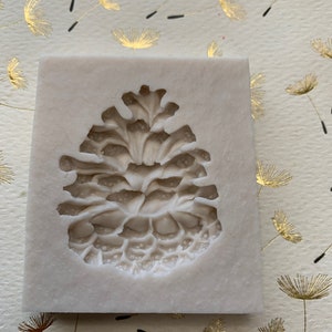 May include: A white silicone mold shaped like a pine cone. The mold has a detailed texture to create a realistic pine cone design.